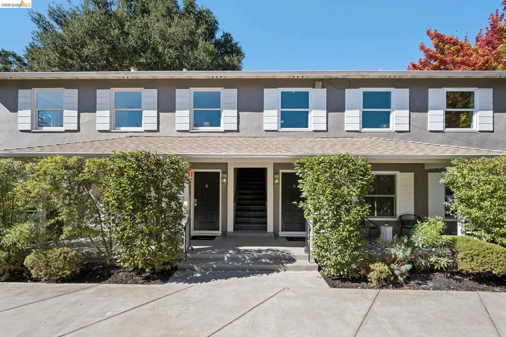 Multi-unit exterior featuring a neutral stucco facade, white shutters, double-hung windows, a concrete entryway, and mature landscaping