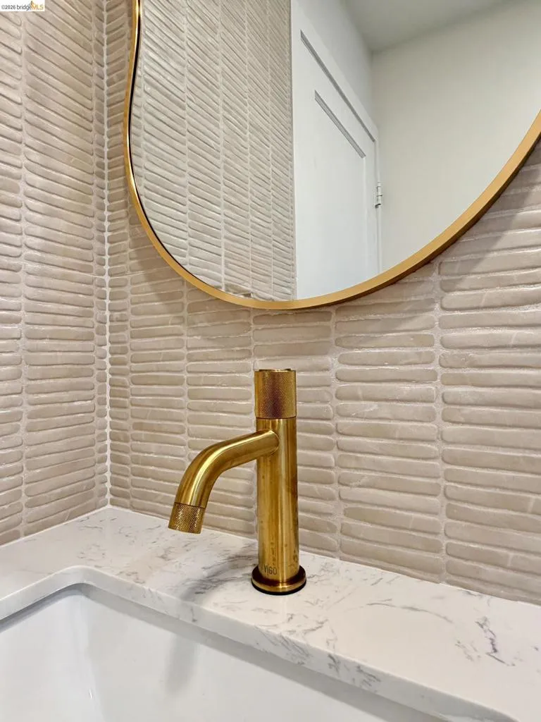 Textured elongated tile backsplash, brass-finish single-handle faucet, oval mirror with brass-tone frame, and solid-surface countertop with undermount​​‌​​​​‌​​‌‌​​‌​​​‌‌​​​‌​​‌‌​​‌‌​​‌‌​​​​ sink