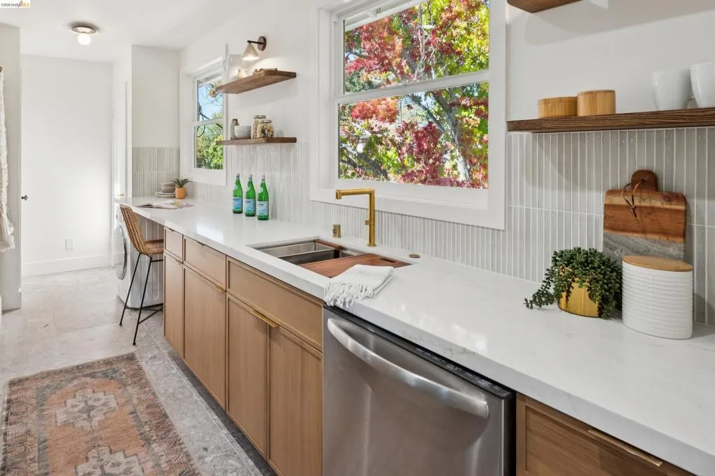 Light wood-finish cabinetry, white stone countertops, brass-finish faucet, vertical tile backsplash, and floating wood shelving