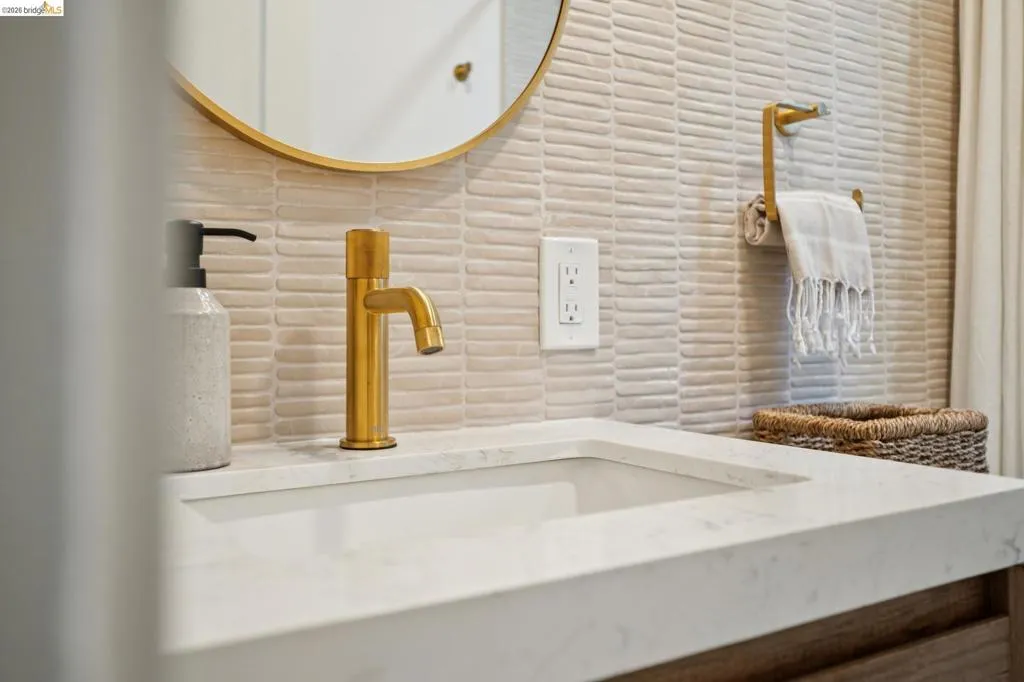 Modern vanity featuring a white composite countertop, integrated rectangular sink, brass-finish faucet, textured horizontal wall tile, and a circular wall-mounted mirror with a gold-tone frame