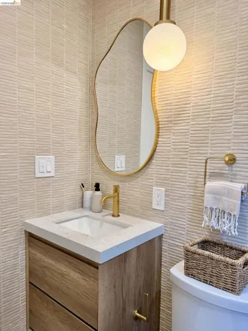 Wall-mounted vanity featuring wood-finish cabinetry and a stone-composite countertop, complemented by a brass faucet, organic-shaped mirror, and globe pendant light set against textured tile walls