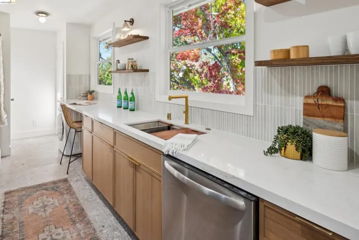 Light wood-finish cabinetry, white stone countertops, brass-finish faucet, vertical tile backsplash, and floating wood shelving