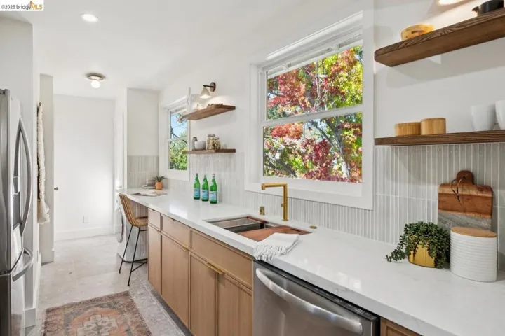 Kitchen featuring light wood-finish cabinetry, quartz countertops, vertical tile backsplash, floating wood shelving, and a brass-finish faucet