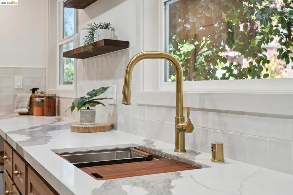 Undermount workstation sink with an integrated cutting board, brass pull-down faucet, stone-patterned countertop, square tile backsplash, and floating wood shelving