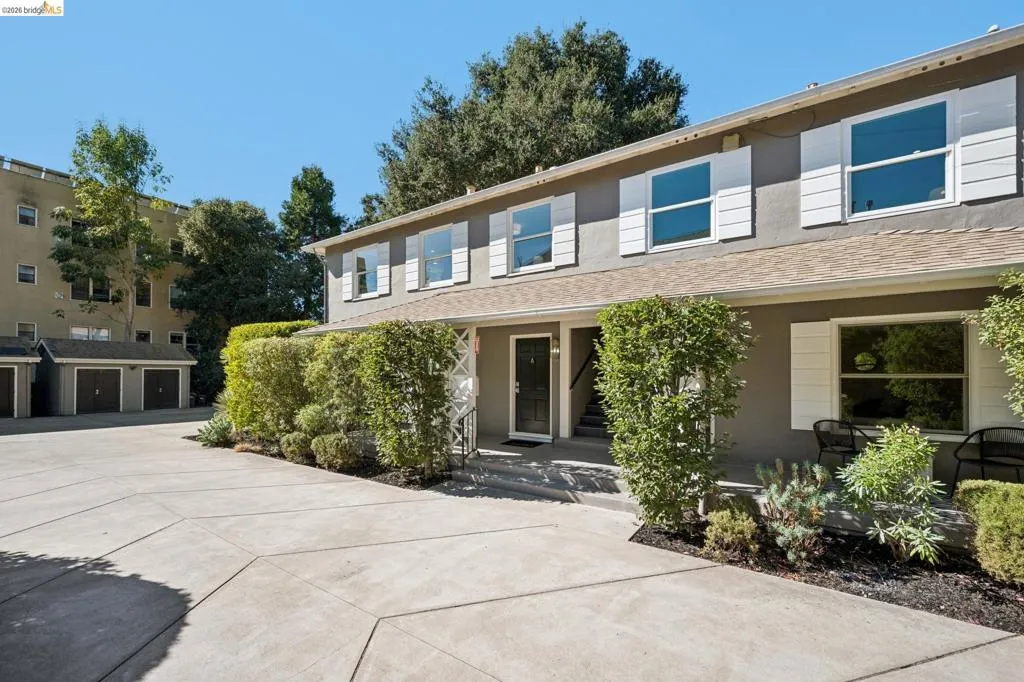 Two-story exterior featuring a gray stucco facade, white shutters, a covered porch, and expansive concrete driveway area