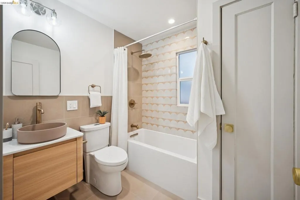 Floating wood-finish vanity with a vessel sink, arched wall mirror, patterned ceramic tile shower surround, and brass-tone plumbing fixtures