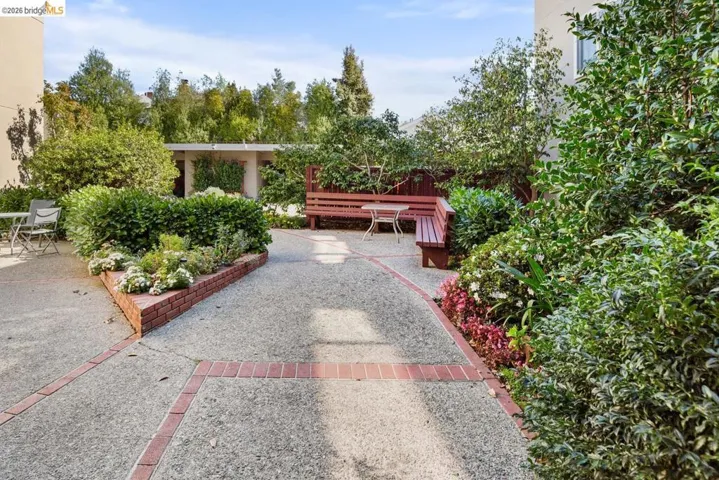Outdoor courtyard featuring exposed aggregate concrete paving, brick-bordered planter beds, built-in wooden bench seating, and mature landscaping