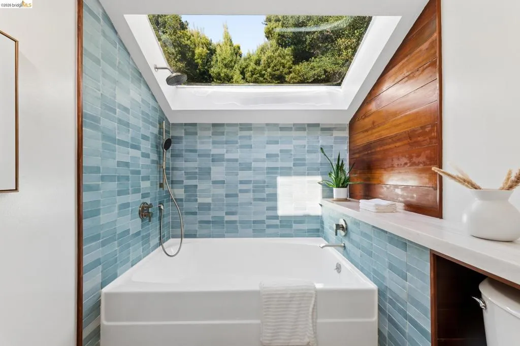Soaking tub featuring a large skylight, blue tiled walls, integrated wood-finish wall paneling, and a polished metal handheld shower fixture