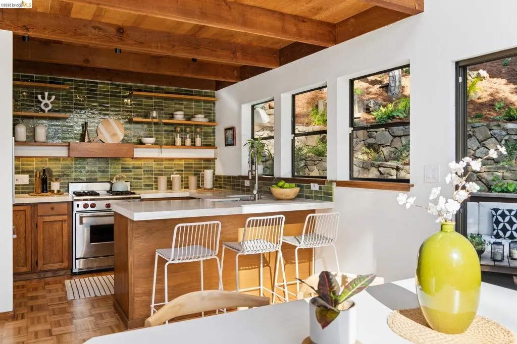 Kitchen featuring exposed wood ceiling beams, green glazed tile backsplash, open wood shelving, wood cabinetry, and a stone retaining wall exterior visible through multiple windows