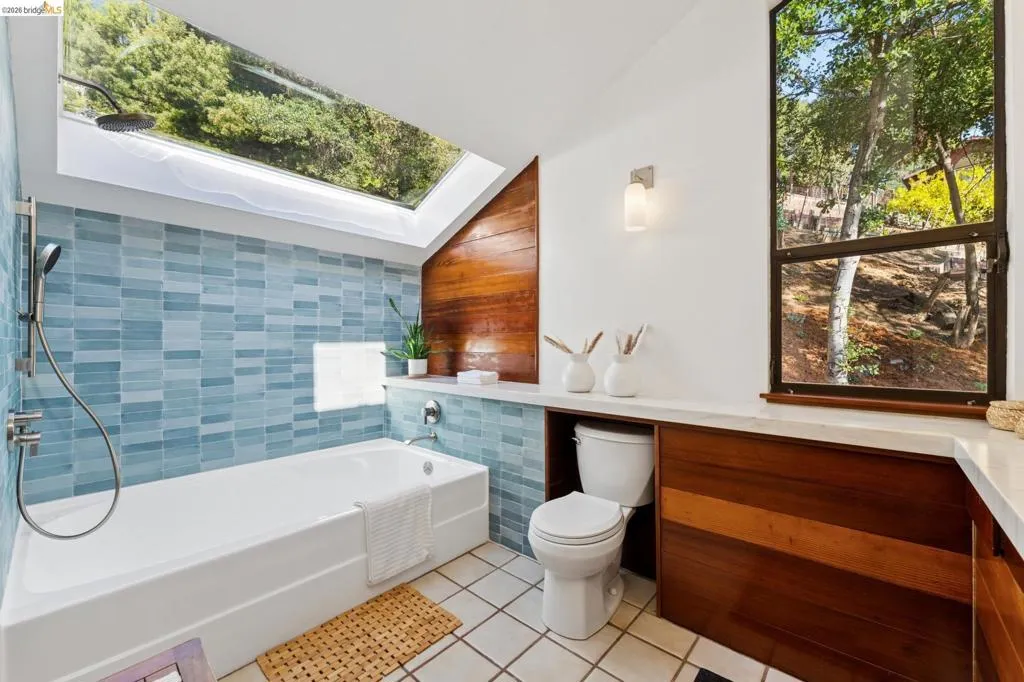 Bathroom featuring a built-in bathtub, blue glass-tiled shower surround, large skylight, natural wood-paneled accents, and a large window