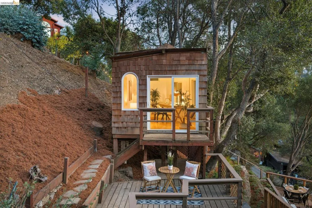 Shingle-sided detached structure featuring a raised wooden deck, arched window, and sliding glass entry door situated on a sloped lot with natural landscaping