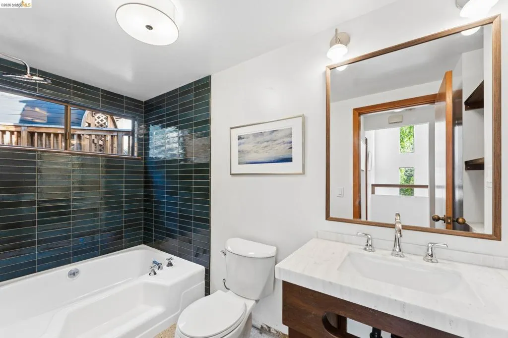 Bathroom featuring a deep soaking tub, dark glass-tile shower surround, stone-top vanity with wood-finish cabinetry, and a large wood-framed mirror