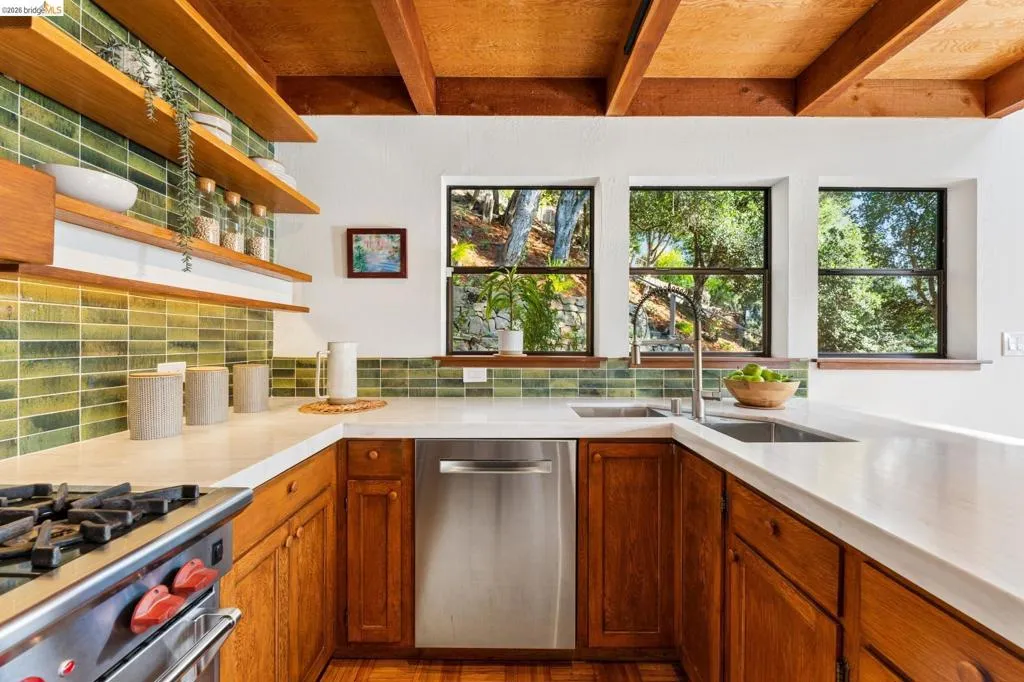 Kitchen featuring wood cabinetry, solid-surface countertops, green tile backsplash, open wood shelving, and exposed ceiling beams