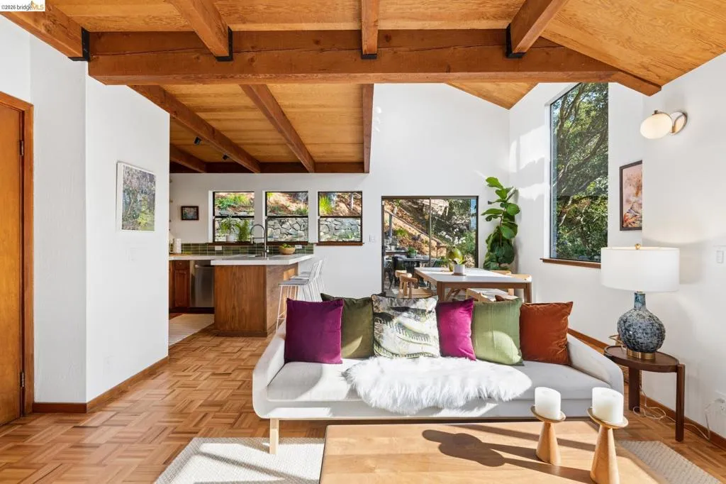 Vaulted ceiling with exposed wood beams, parquet wood flooring, large floor-to-ceiling windows, and wood-finish kitchen cabinetry