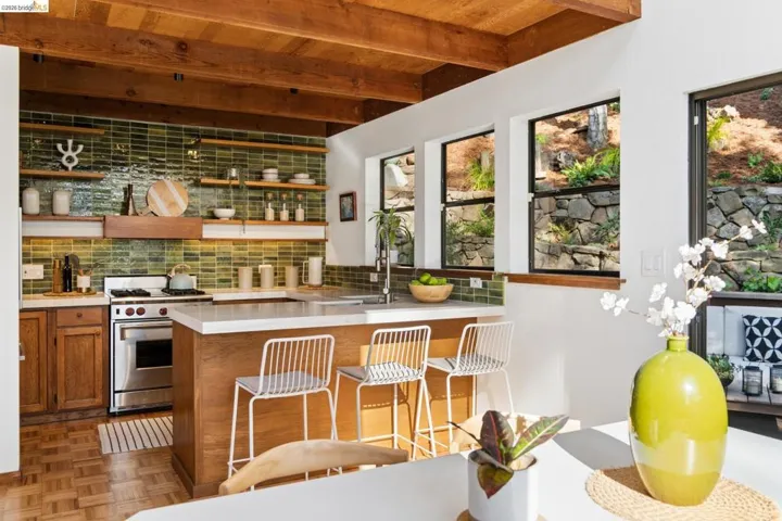 Kitchen featuring exposed wood ceiling beams, green glazed tile backsplash, open wood shelving, wood cabinetry, and a stone retaining wall exterior visible through multiple windows