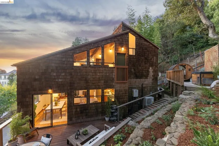 Exterior featuring dark cedar shake siding, expansive tiered wooden deck, geometric multi-pane windows, and stone-retaining wall landscaping