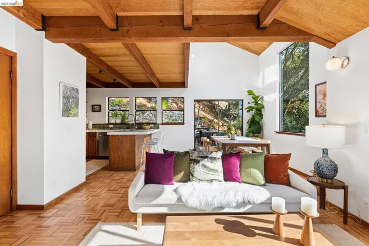 Vaulted ceiling with exposed wood beams, parquet wood flooring, large floor-to-ceiling windows, and wood-finish kitchen cabinetry
