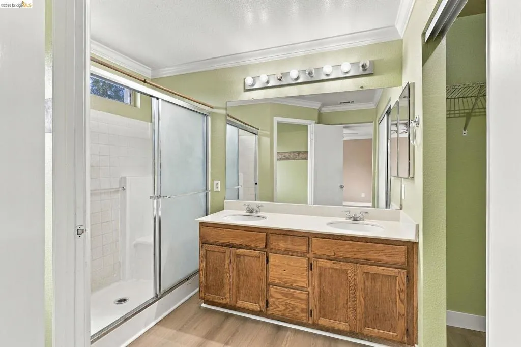 Dual-sink vanity with wood-finish cabinetry, solid-surface countertop, full-width mirror, glass-enclosed shower, and wood-finish flooring