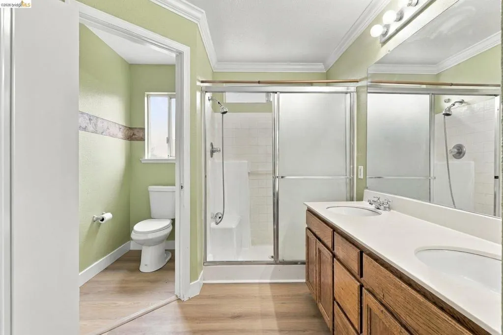 Dual-sink vanity with a solid-surface countertop, wood-finish cabinetry, walk-in shower with a sliding glass enclosure, and wood-finish flooring