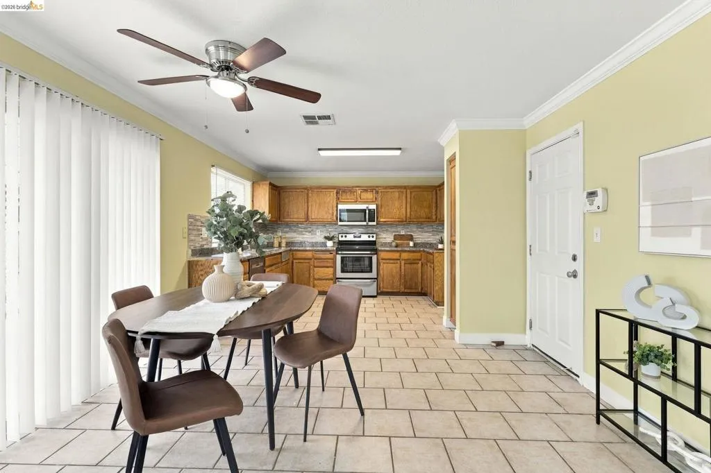 Kitchen featuring wood cabinetry, stone-finish backsplash, neutral tile flooring, crown molding, and a ceiling fan with integrated lighting