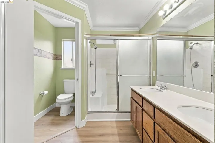 Dual-sink vanity with a solid-surface countertop, wood-finish cabinetry, walk-in shower with a sliding glass enclosure, and wood-finish flooring