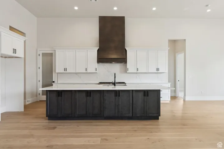 Kitchen with custom exhaust hood, light wood-style floors, a center island with sink, light countertops, and white cabinetry