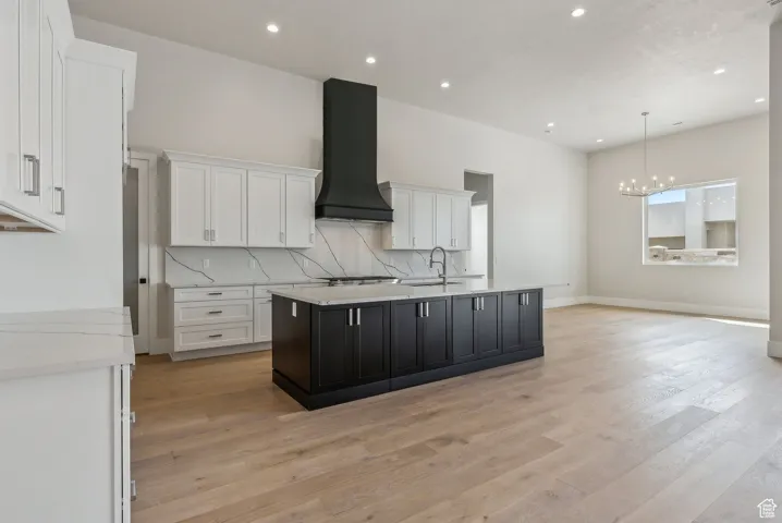 Kitchen featuring custom exhaust hood, tasteful backsplash, white cabinets, a center island with sink, and a sink