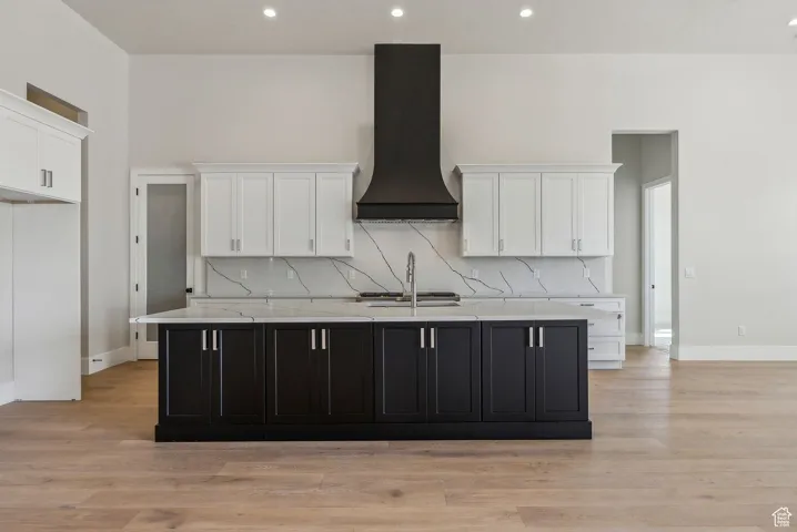 Kitchen with custom exhaust hood, a kitchen island with sink, white cabinetry, light wood finished floors, and backsplash