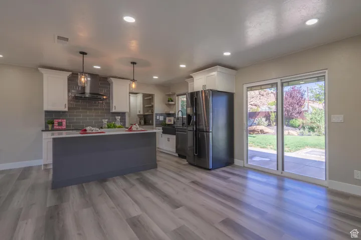 Kitchen with pendant lighting, light hardwood / wood-style floors, white cabinetry, wall chimney range hood, and stainless steel fridge
