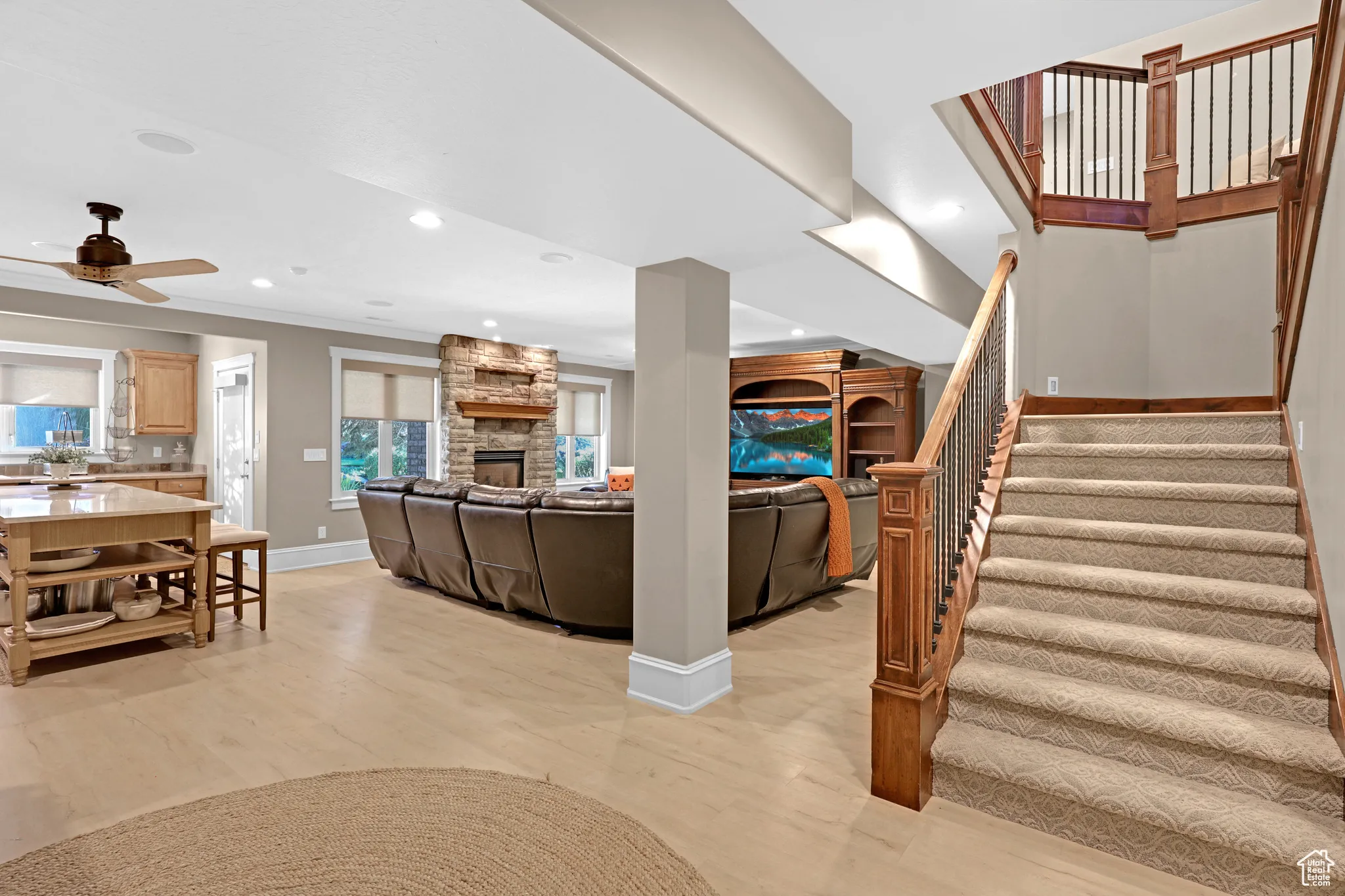 Downstairs living room featuring a stone fireplace, ornamental molding, light wood-type flooring, and ceiling fan