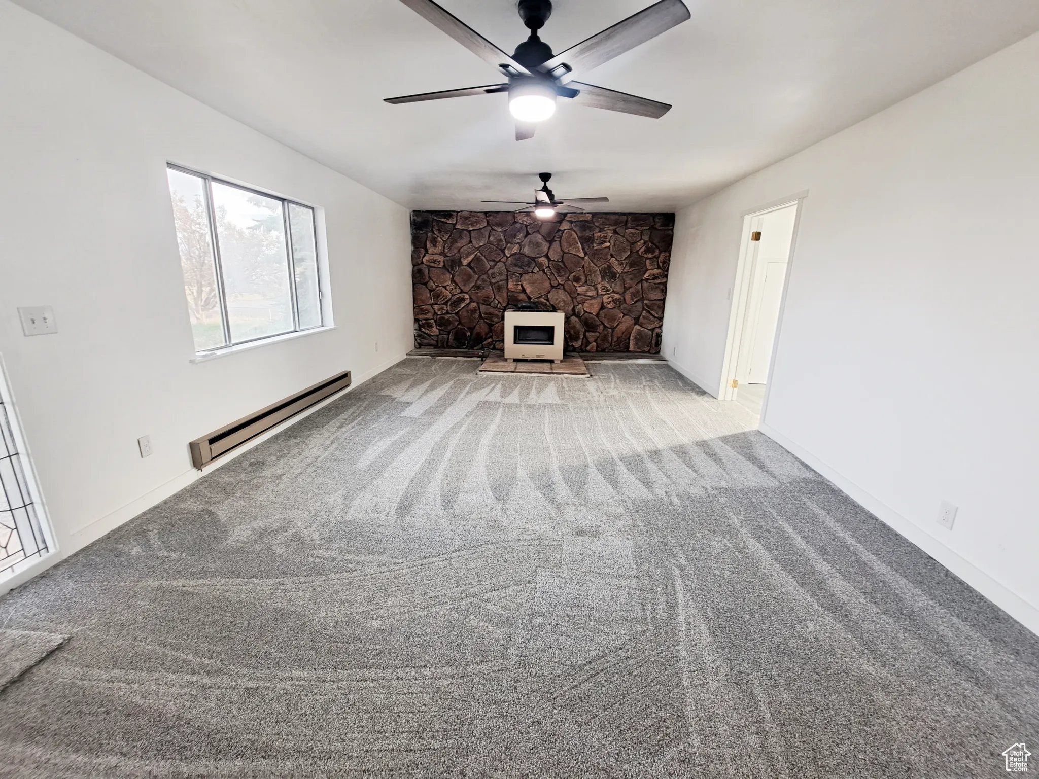 Living room with baseboard heating, New light colored carpet, ceiling fan, New paint, and a stone propane fireplace