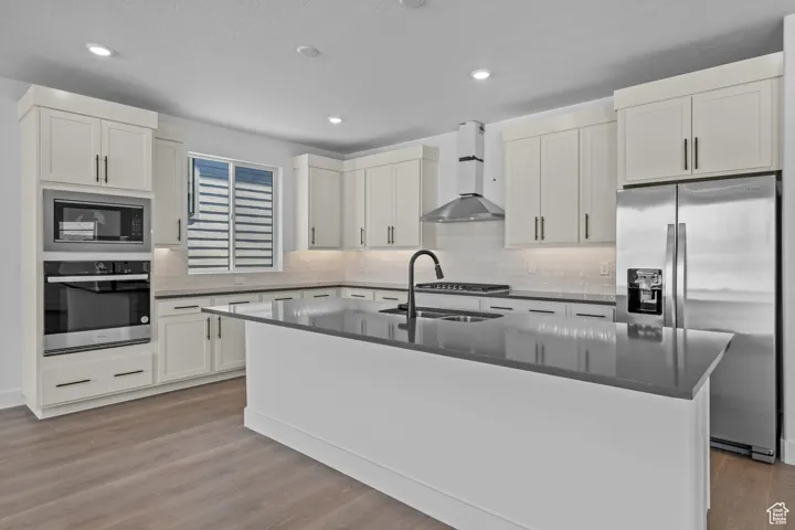 Kitchen featuring appliances with stainless steel finishes, sink, wall chimney range hood, a center island with sink, and light hardwood / wood-style flooring