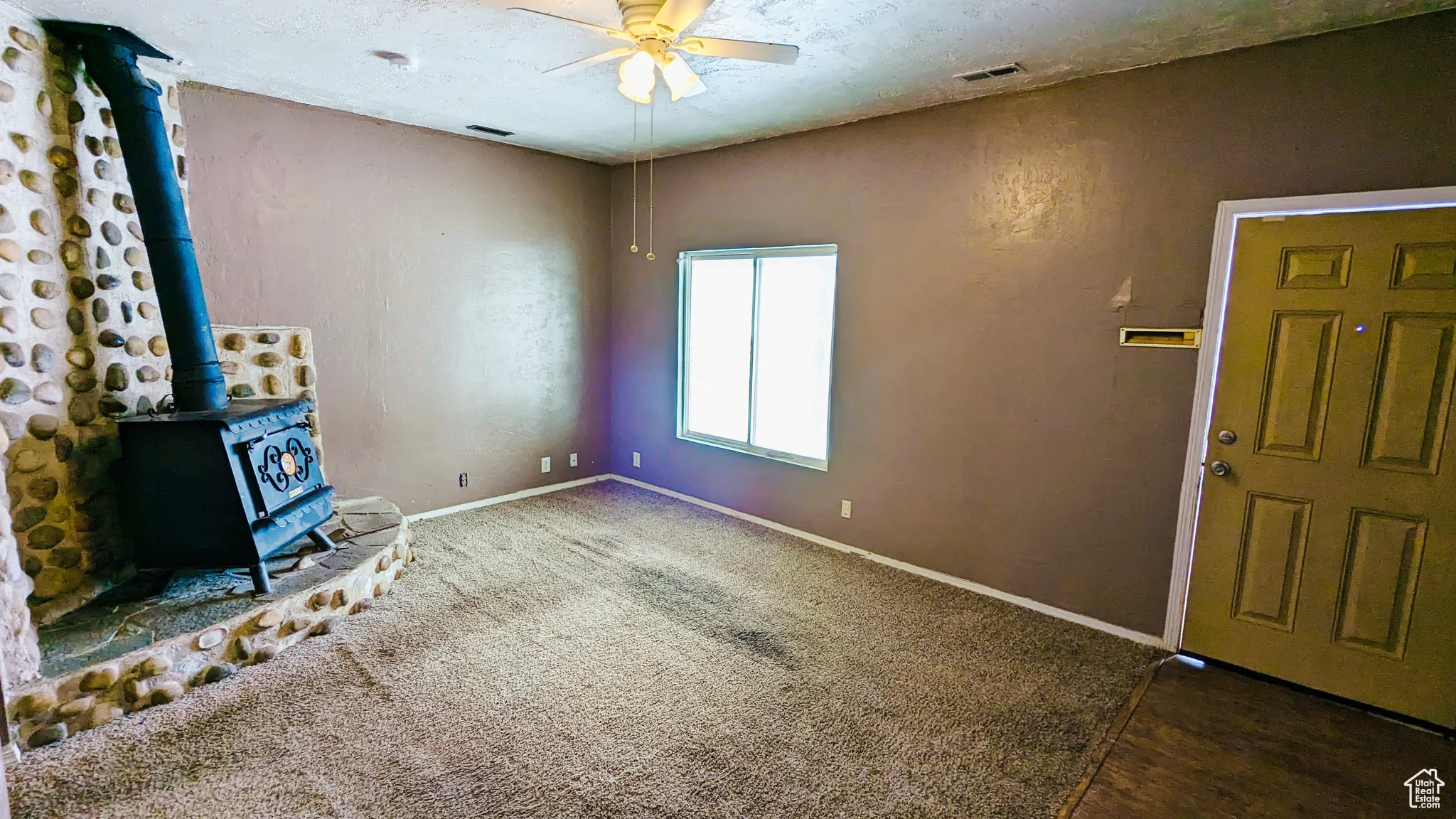 Unfurnished living room featuring dark colored carpet, a wood stove, and ceiling fan