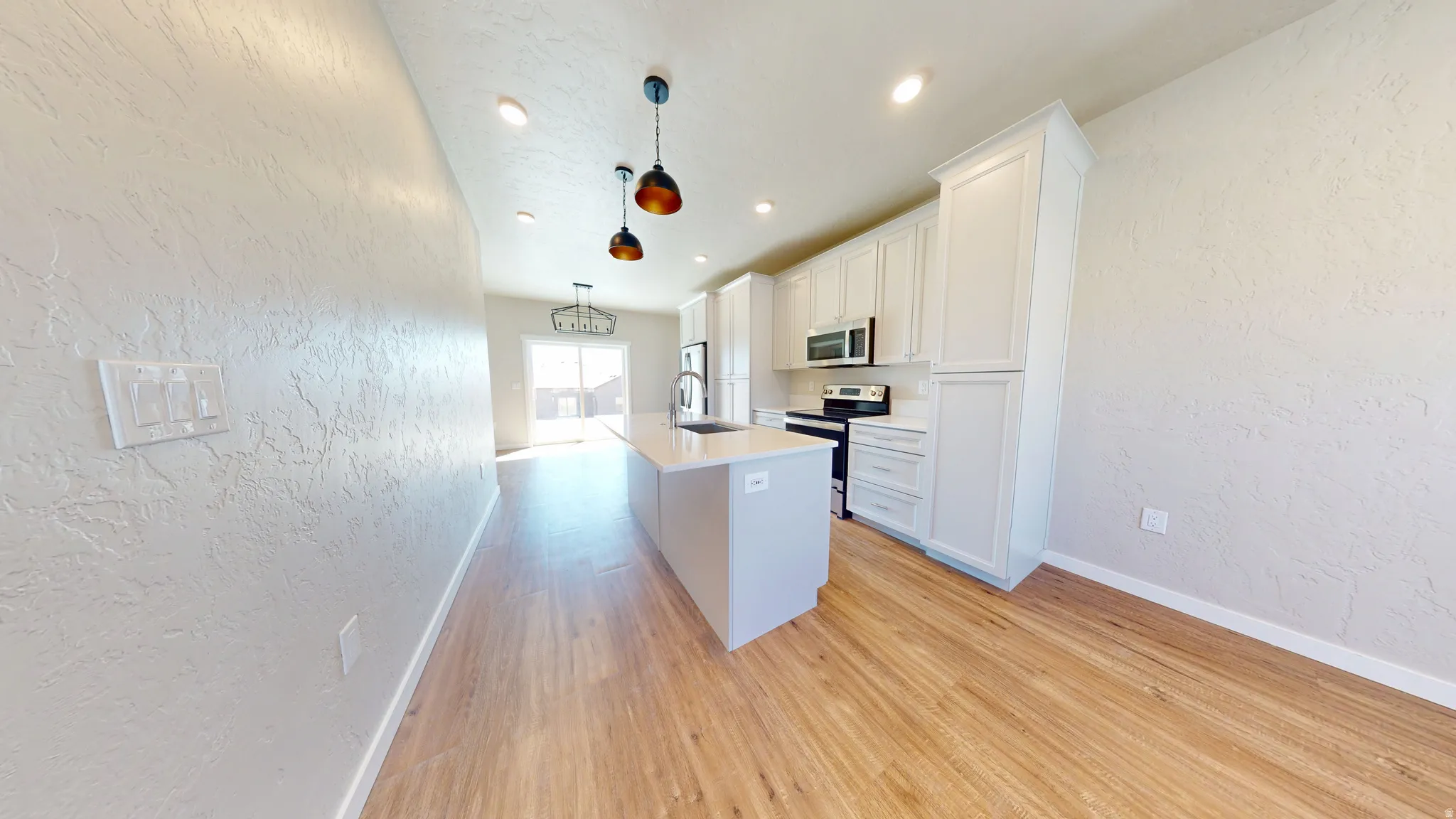Kitchen with a textured wall, an island with sink, appliances with stainless steel finishes, white cabinetry, and light wood-type flooring