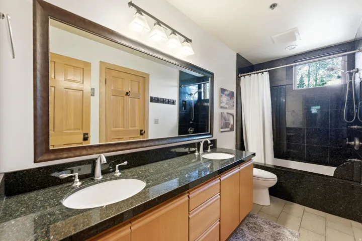 Bathroom featuring a dual vanity with a dark speckled countertop, light wood-finish cabinetry, and a large framed mirror