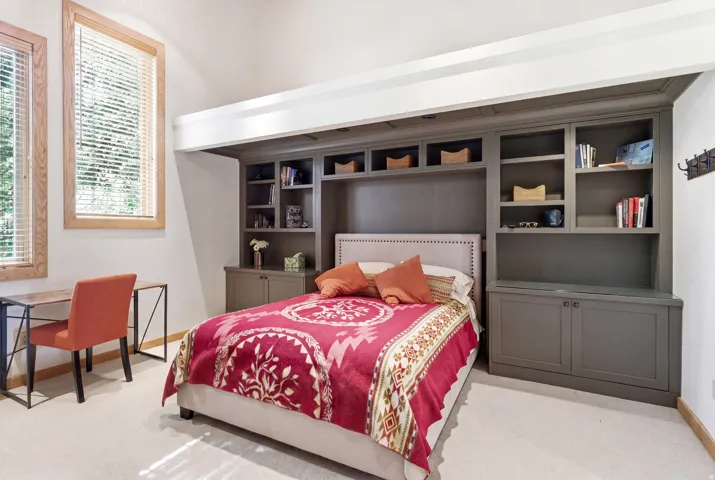 Built-in cabinetry with shelving and storage, two natural wood-framed windows with blinds, and light-toned carpet flooring