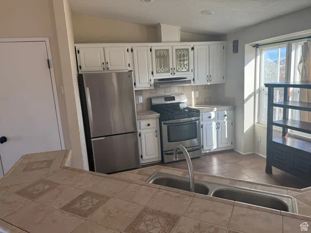 Kitchen with vaulted ceiling, tile countertops, tasteful backsplash, white cabinets, and stainless steel appliances