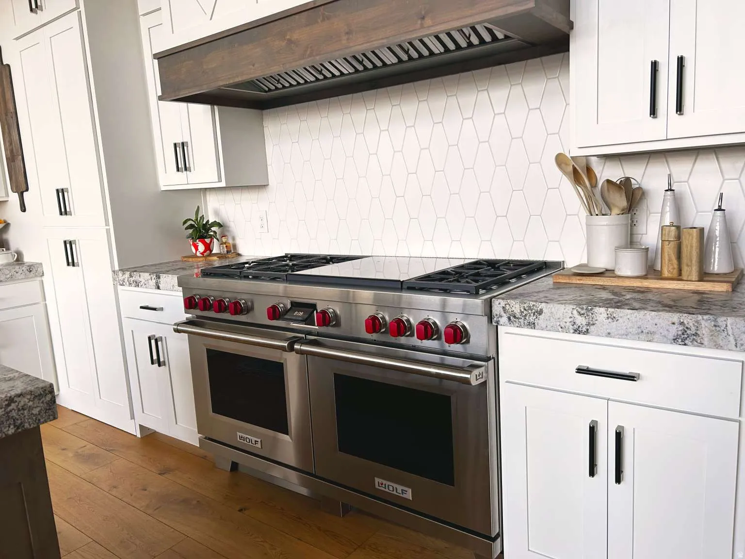 Stainless steel range with a double oven and red control knobs, white shaker-style cabinetry with black pulls, wood-finish flooring, stone-finish countertops, and a white geometric tile backsplash