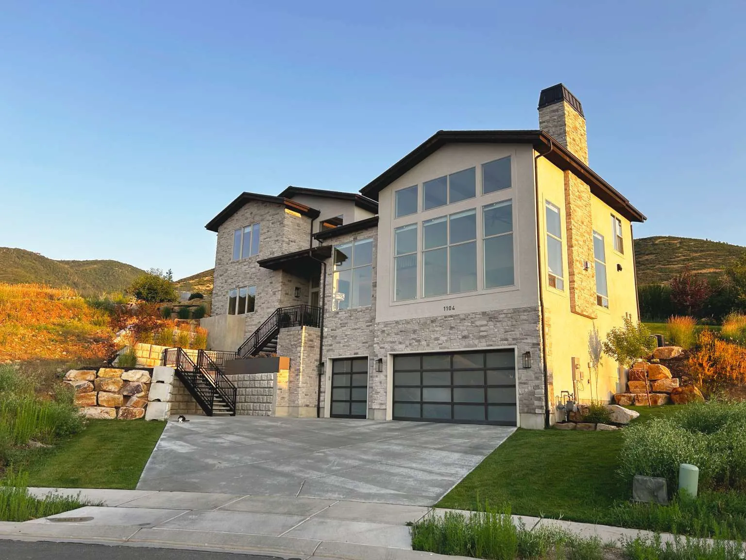 Contemporary multi-level exterior featuring a stucco and stone facade, large format windows, a stone chimney, and an integrated two-car garage with modern doors