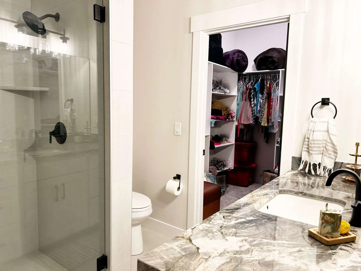 Bathroom featuring a glass-enclosed shower with matte black fixtures, a single vanity with a patterned stone countertop and integrated sink, a toilet, and an organized closet with shelving and hanging space