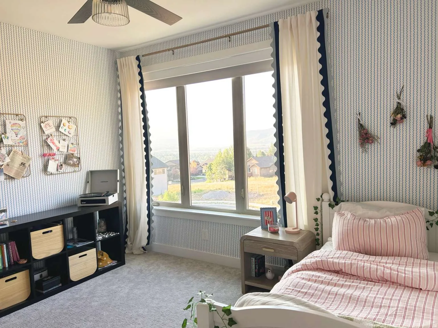 Carpeted room featuring a large window with white window treatments, a ceiling fan with integrated lighting, and patterned wallpaper