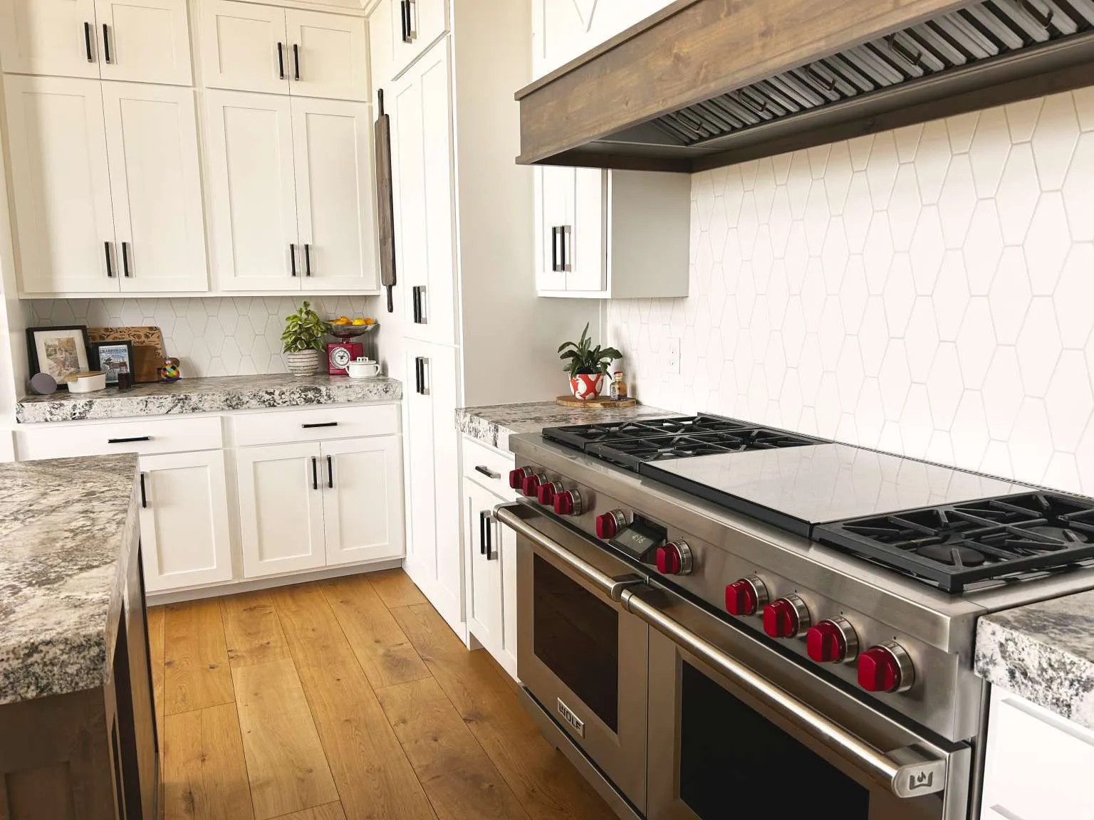 Gourmet kitchen featuring a professional-grade stainless steel range with red knobs, white hexagonal tile backsplash, light-toned wood-finish flooring, white shaker cabinetry with dark hardware, and speckled countertops