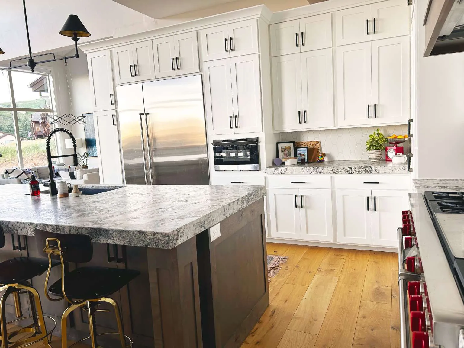 Gourmet kitchen featuring a large central island with integrated sink, white shaker-style cabinetry with dark hardware, light-colored stone-finish countertops, stainless steel appliances, and wood-finish flooring