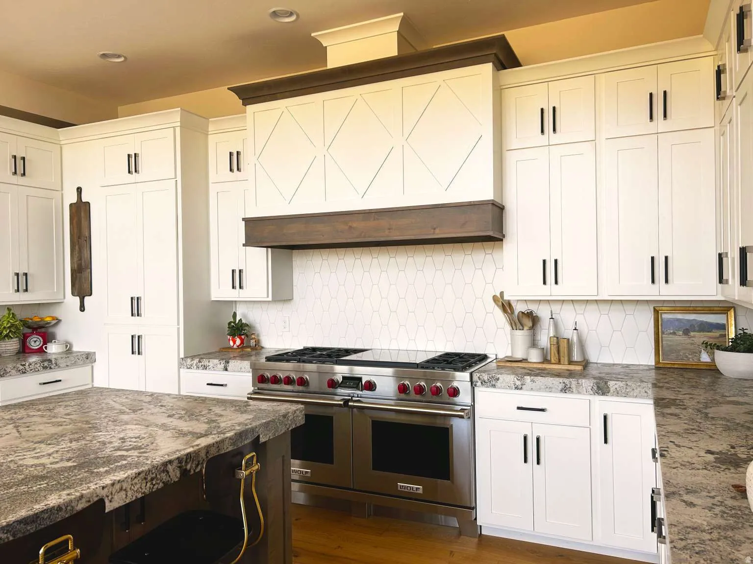 Expansive kitchen featuring a professional-grade stainless steel range with red knobs, a custom wood-trimmed range hood with geometric paneling, and white shaker-style cabinetry