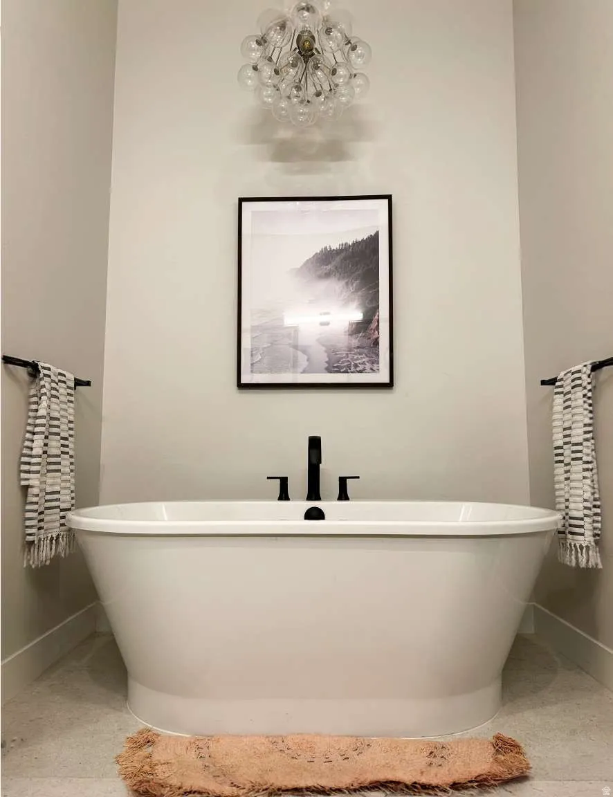 Freestanding soaking tub featuring matte black fixtures, an overhead globe chandelier, light-toned wall paint, and gray tile flooring
