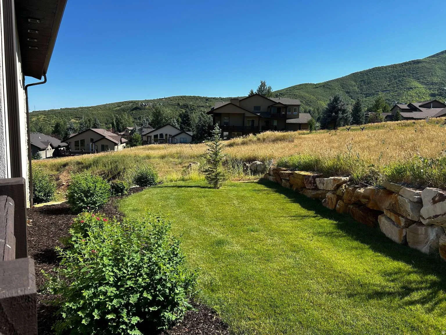 Expansive green lawn framed by a natural stone retaining wall and lush landscaping, featuring a backdrop of distant mountains and residential properties