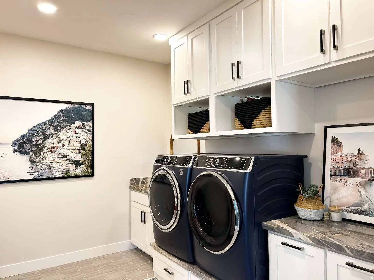 Dedicated laundry room featuring side-by-side front-load washers and dryers in navy blue, white shaker-style cabinetry with brushed black hardware, open shelving with woven storage baskets, quartz countertop workspace, recessed lighting, and framed artwor