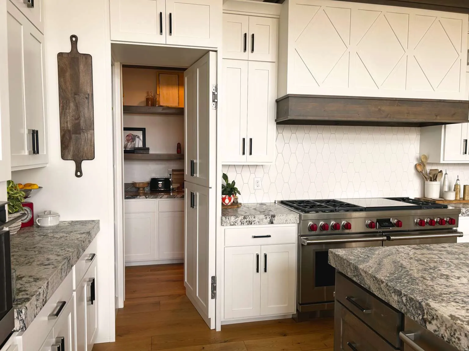 Gourmet kitchen featuring a stainless steel range with red accents, hexagon tile backsplash, granite countertops, custom range hood with wood trim, and a hidden pantry with built-in shelving