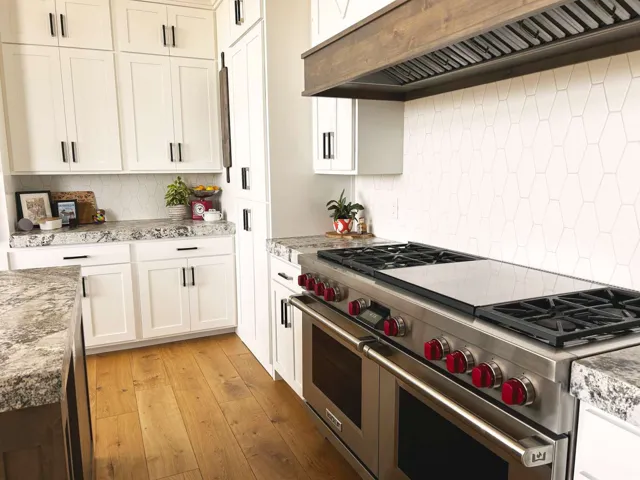 Gourmet kitchen featuring a professional-grade stainless steel range with red knobs, white hexagonal tile backsplash, light-toned wood-finish flooring, white shaker cabinetry with dark hardware, and speckled countertops