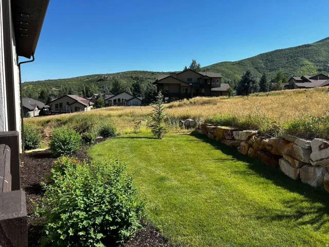 Expansive green lawn framed by a natural stone retaining wall and lush landscaping, featuring a backdrop of distant mountains and residential properties